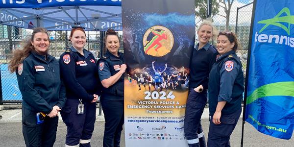 Five female paramedics are standing beside a tennis court while holding on to a standee banner promoting the games.